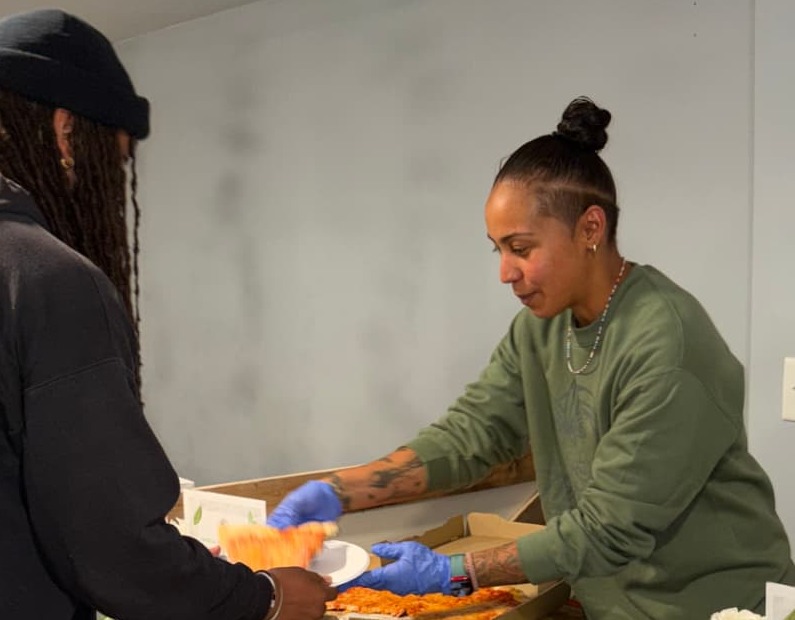 A community member serving a slice of pizza to someone at a shared food table.